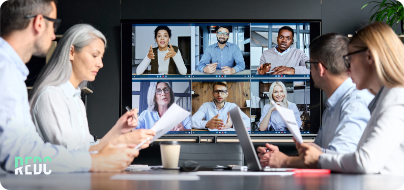 A group in a meeting room in a video conference with people on a screen.
